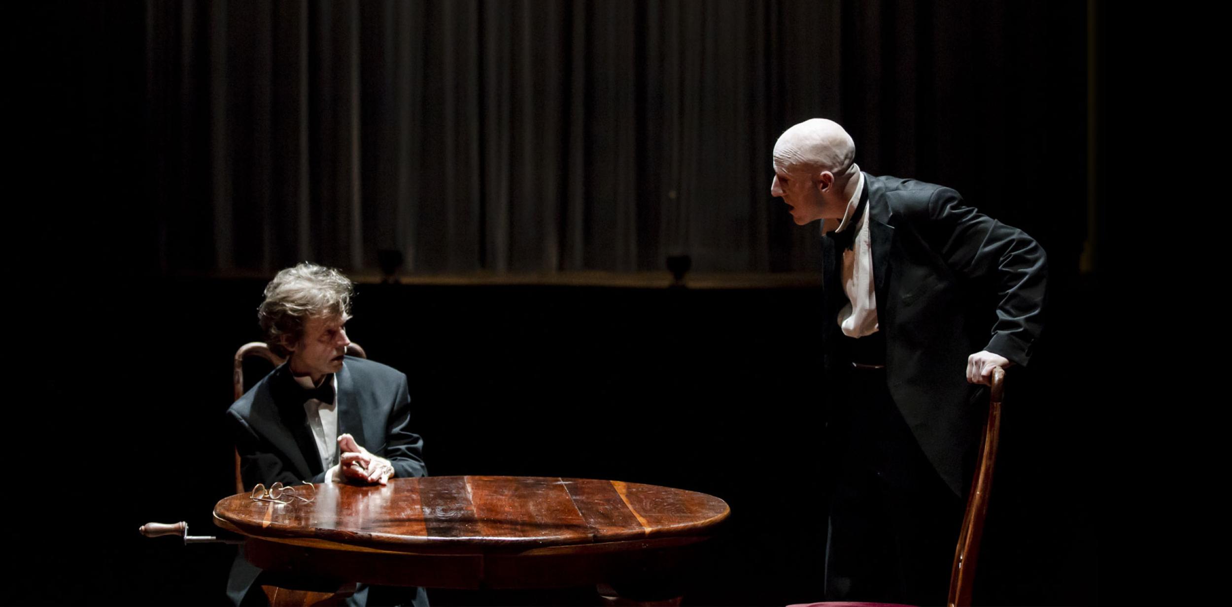 two men in black tie, one sat at a round table, the other standing talking down to him