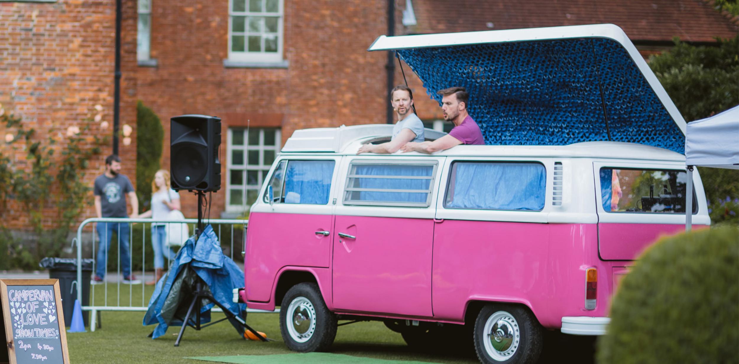 Two men poking their head out the top of a bright pink caravan