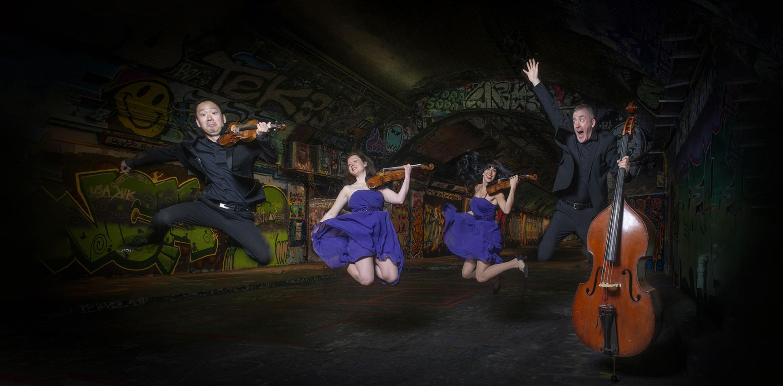 Four classical musicians leaping into the air inside a tunnel covered in graffiti