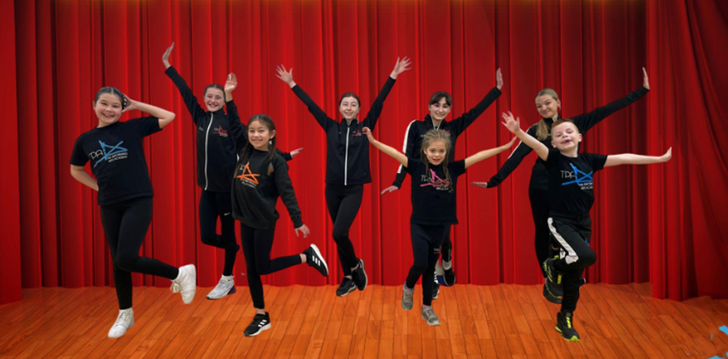 8 young people in black jumping and looking happy in front of a red stage curtain and title High School The Musical Edited