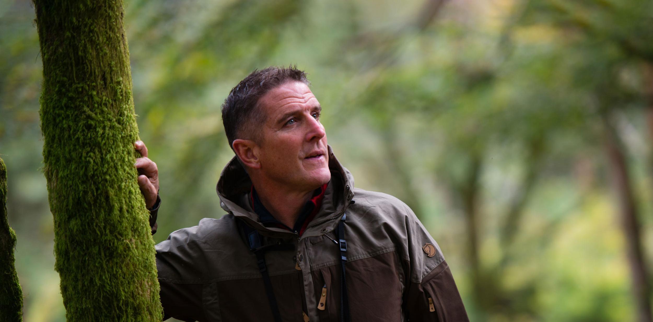 Iolo Williams leaning against a tree in a forrest.