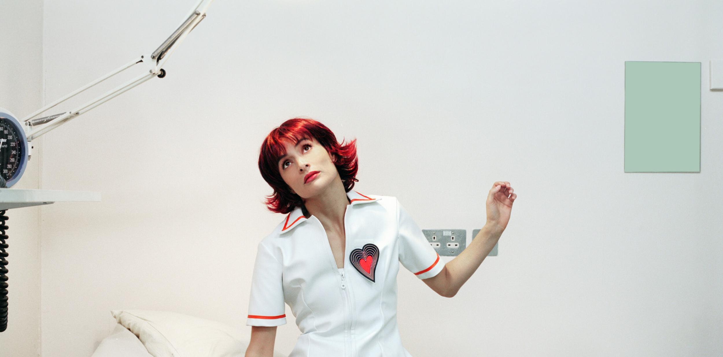 A person with red hair in a nurses outfit sitting on a doctors table looking curiously upward.