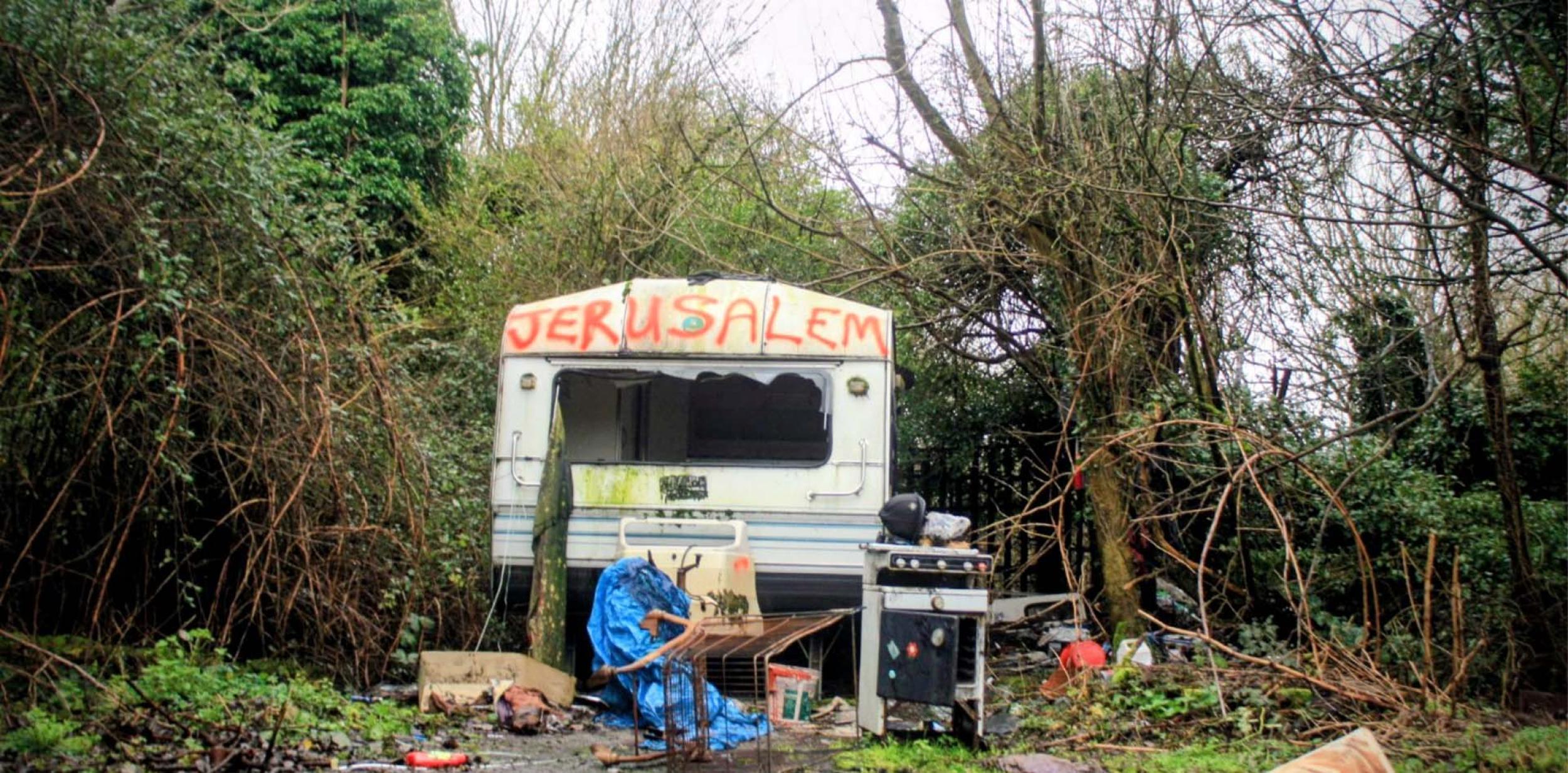 A beat-up-looking caravan surrounded by shrubbery with Jerusalem spray-painted on to it. 