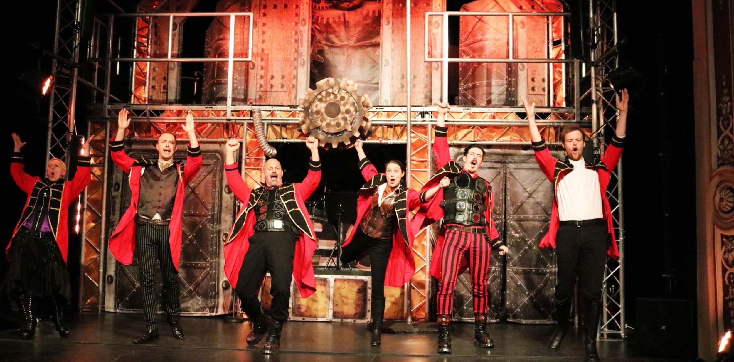 Six actors in red and black uniforms on stage looking animated