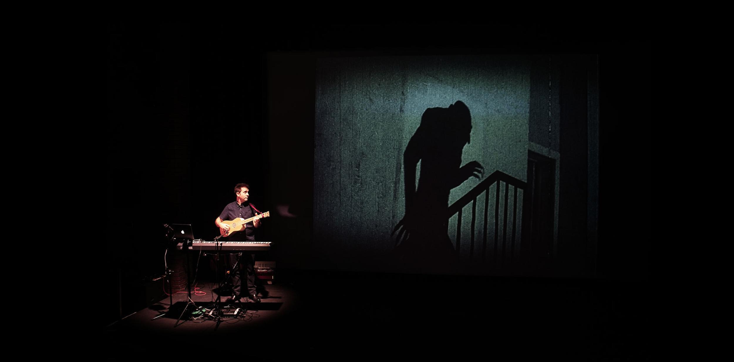 A person playing guitar and keyboard, next to a large cinema screening
