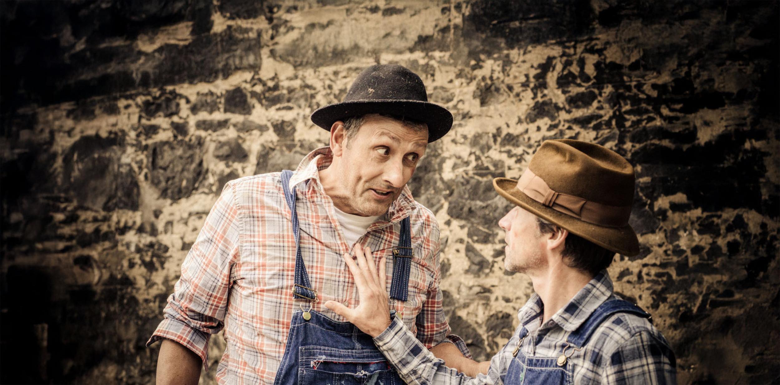 Two men, one tall and the other short, dressed like farmers, talking in front of a stone wall.