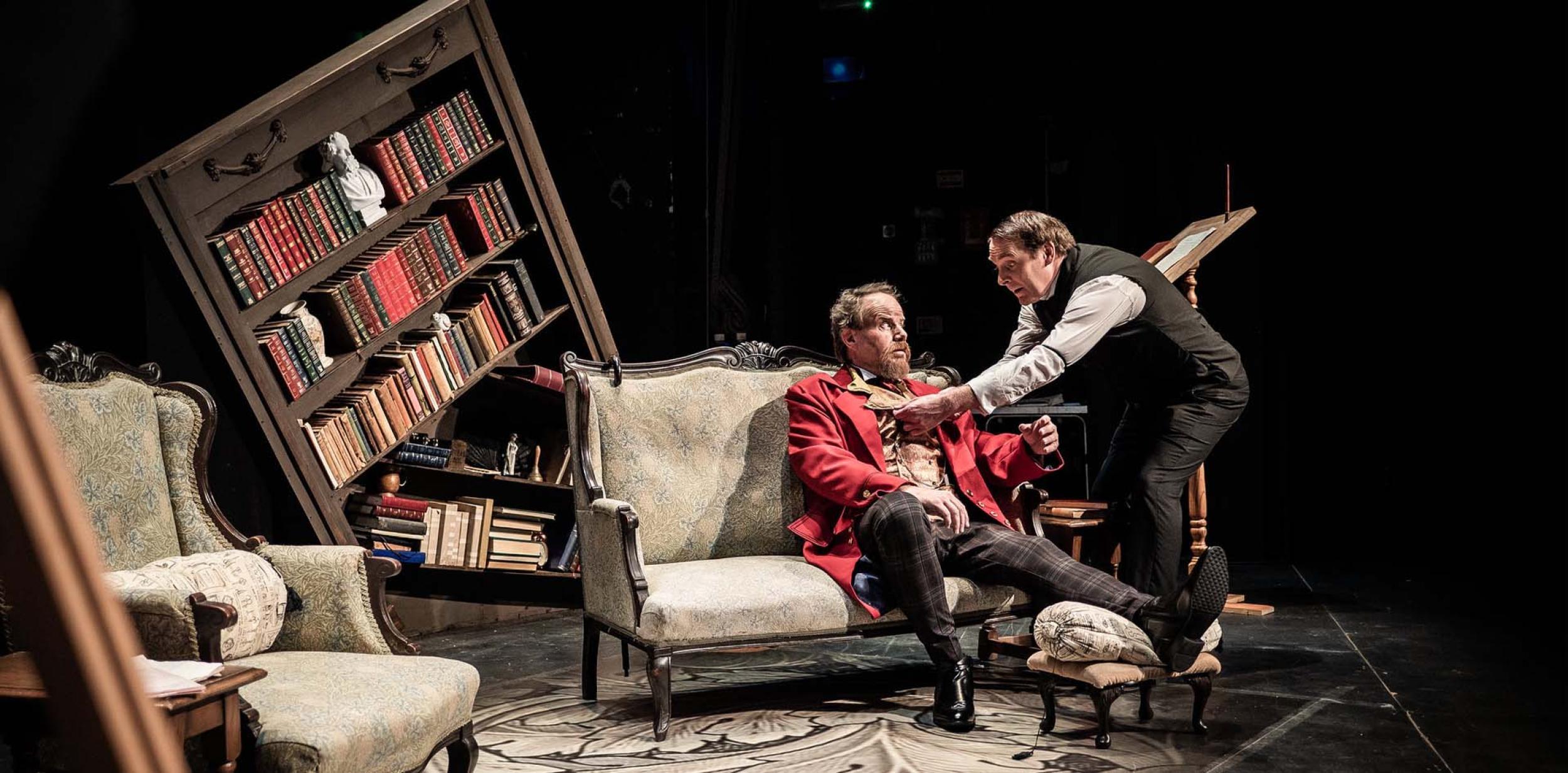 Two actors on stage, one on a sofa the other standing, in front of a wonky bookcase