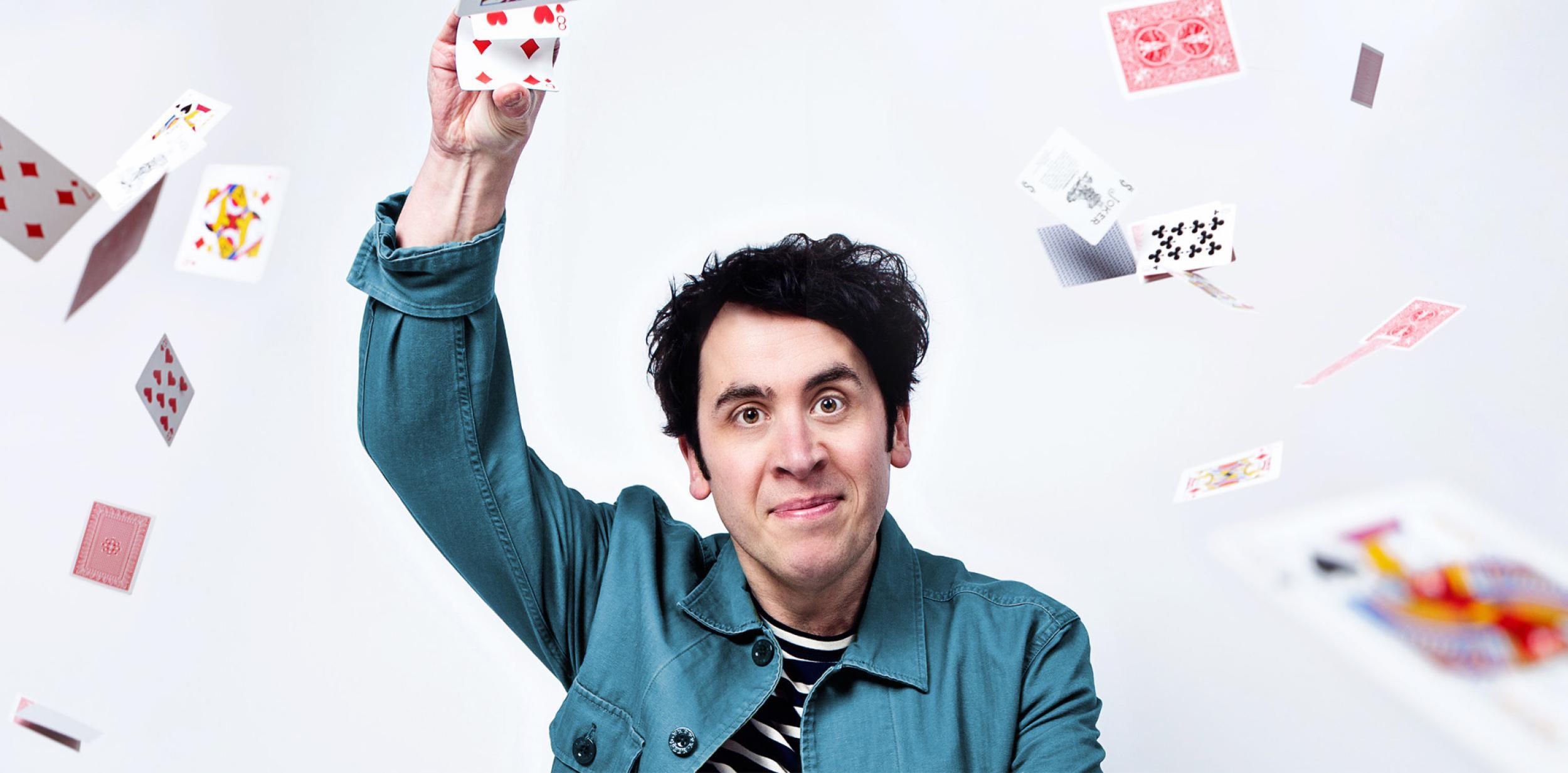 Man in denim shirt with arm raised holding playing card, with other playing cards flying around the space
