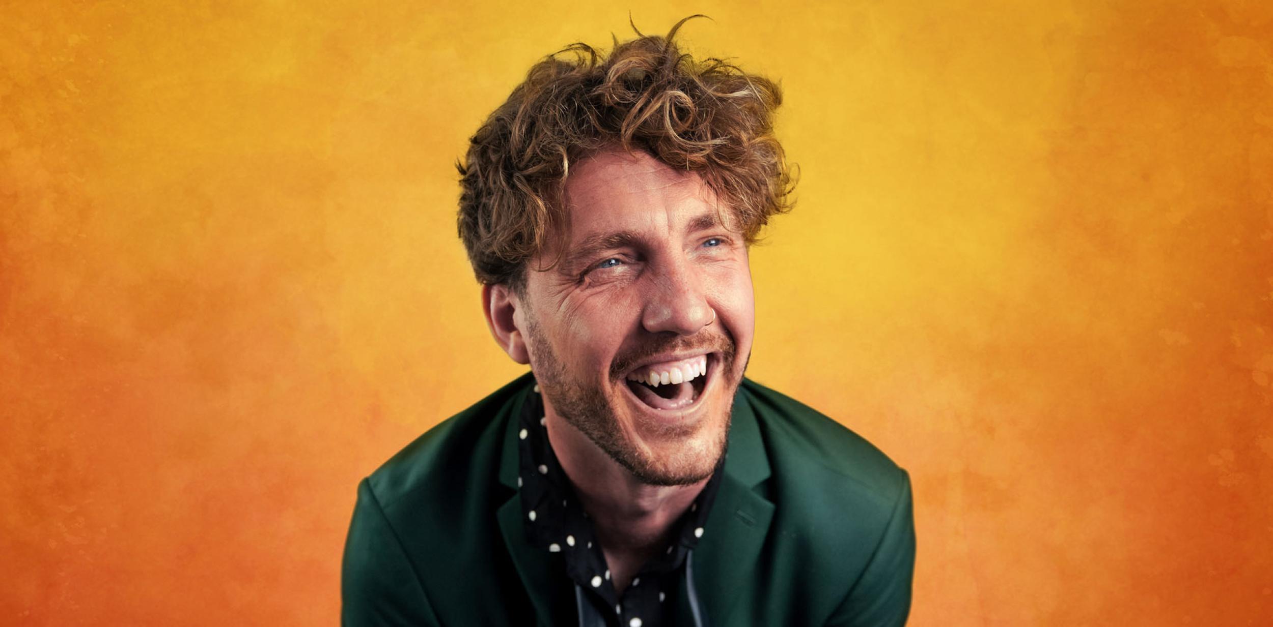 Man with curly hair laughing looking to the right, in a dark green informal suit with a yellow background