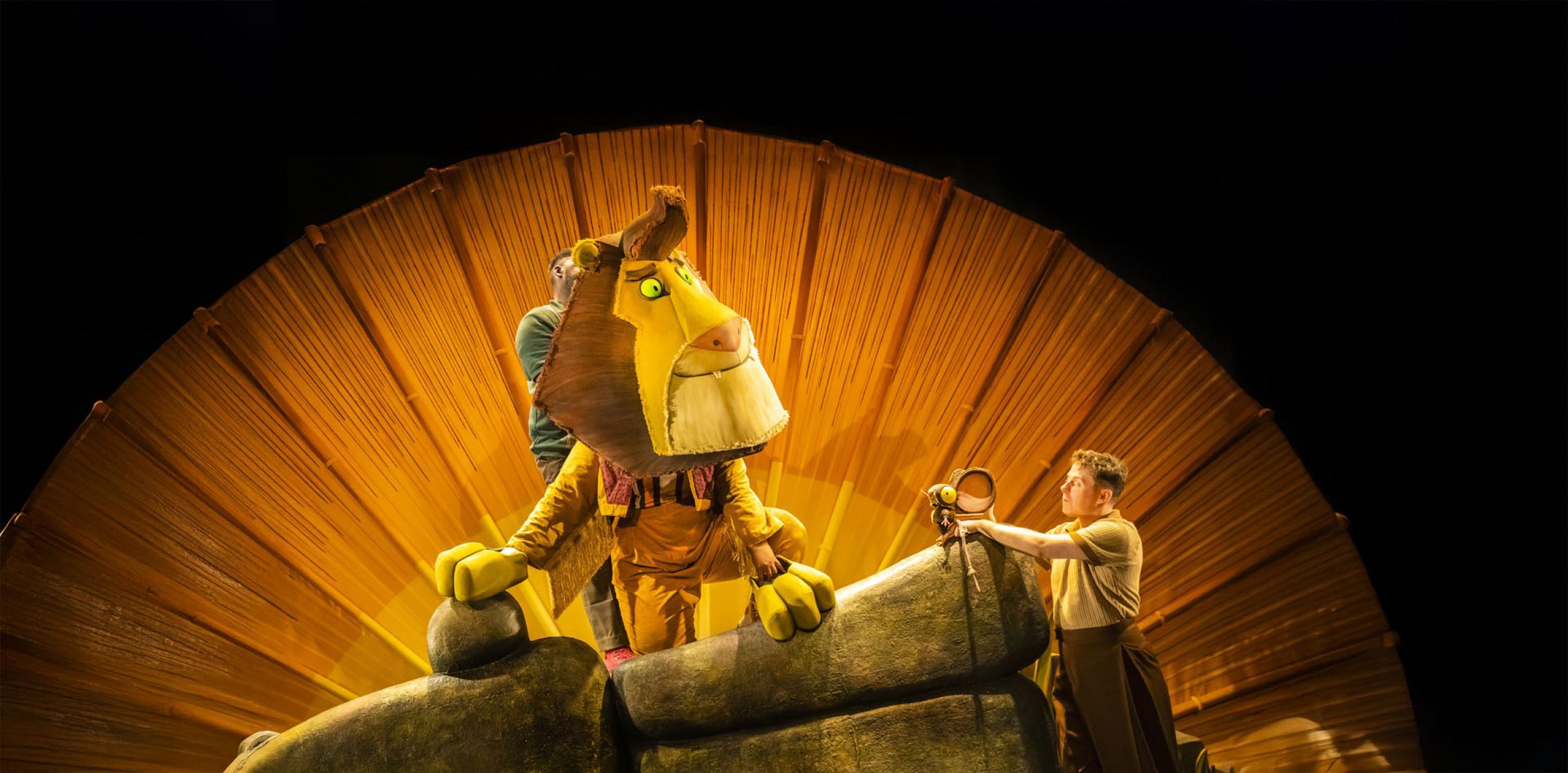 A performer dressed as a lion lounging on a rock with the sun in the backdrop.