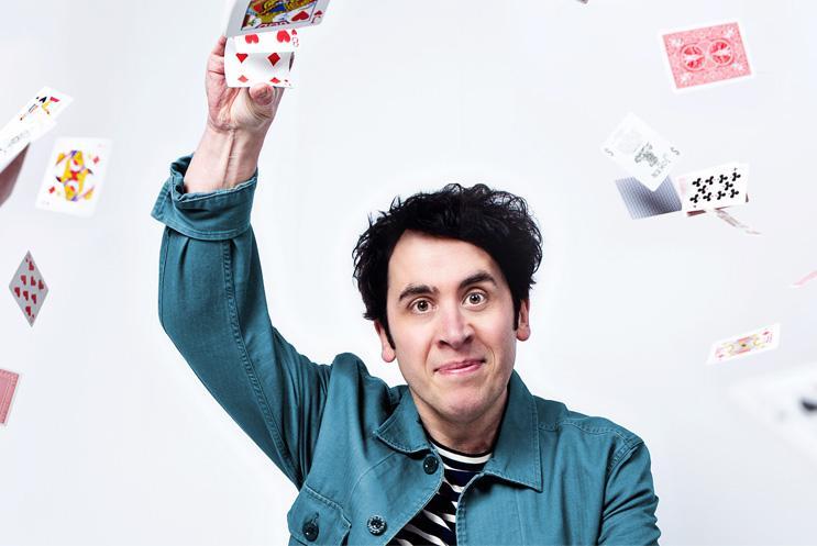 Man in denim shirt with arm raised holding playing card, with other playing cards flying around the space
