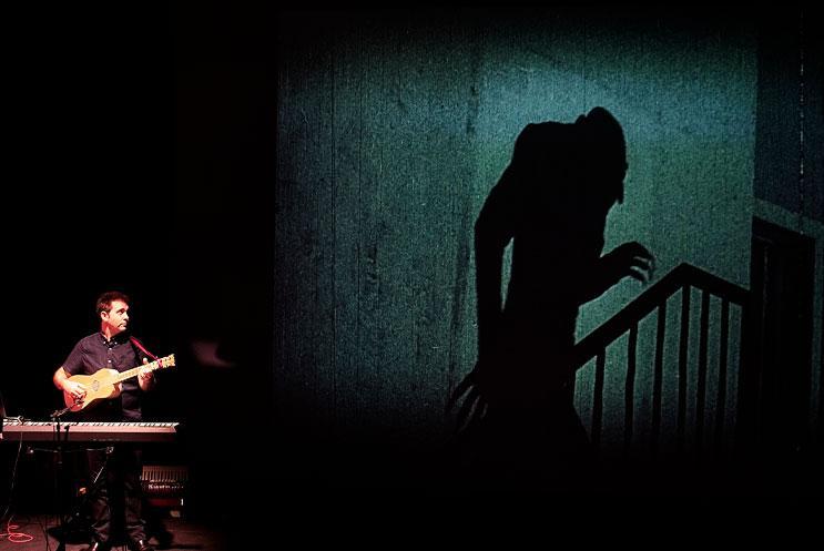 A person playing guitar and keyboard, next to a large cinema screening