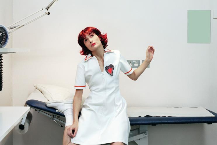 A person with red hair in a nurses outfit sitting on a doctors table looking curiously upward.