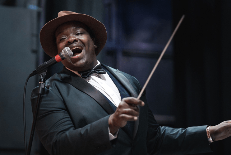 Marvin dressed in a suit and bow tie, singing into a mic and conducting.