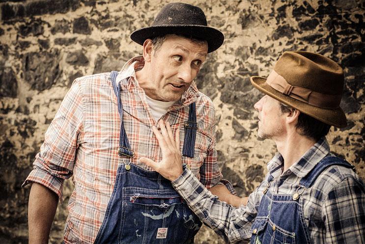 Two men, one tall and the other short, dressed like farmers, talking in front of a stone wall.