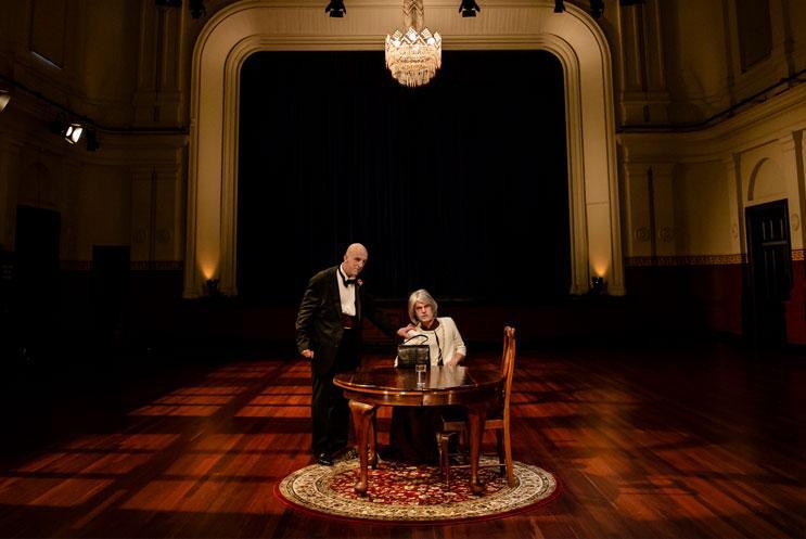 Woman sat at a table with a man stood next to her in an empty old music hall