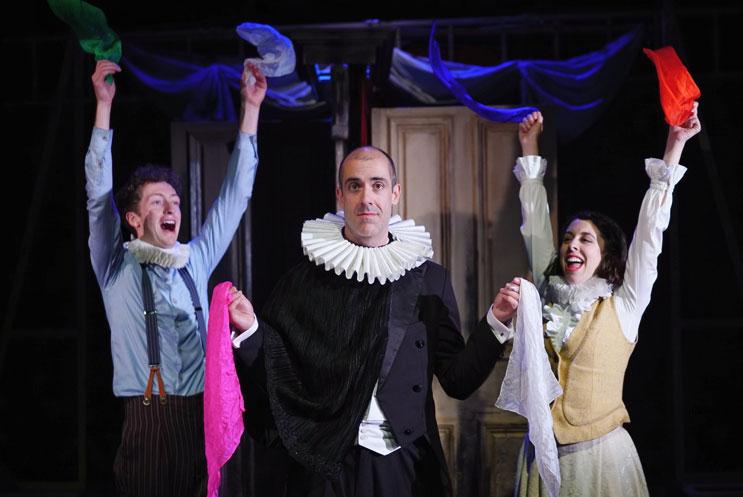 Actor wearing ruff with hankerchiefs plus two actors behind with their arms in the air