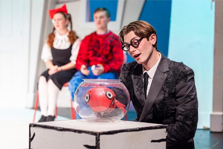 A performer in a suit puppeteering a goldfish in a fishbowl.