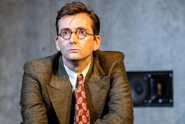 David Tennant in a suit and wearing glasses. He's bending down and looking past the camera.