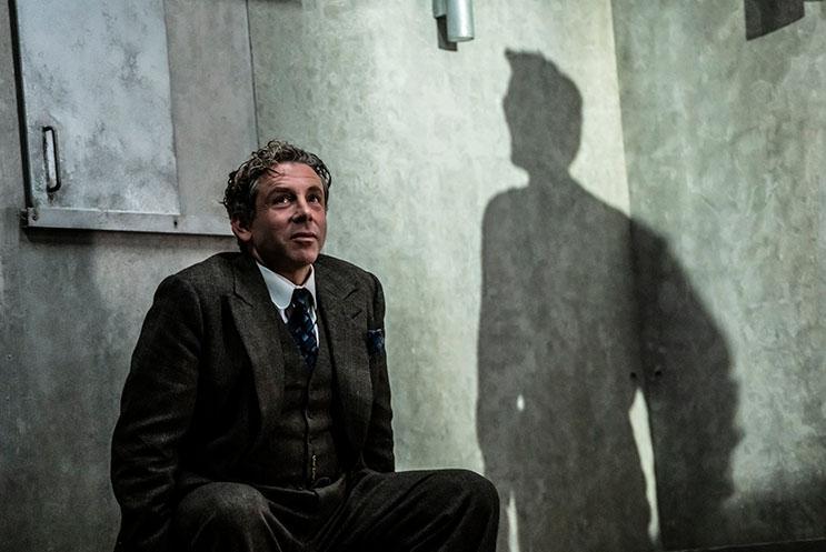 Elliot Levey is sat against a wall, a looming shadow beside him.