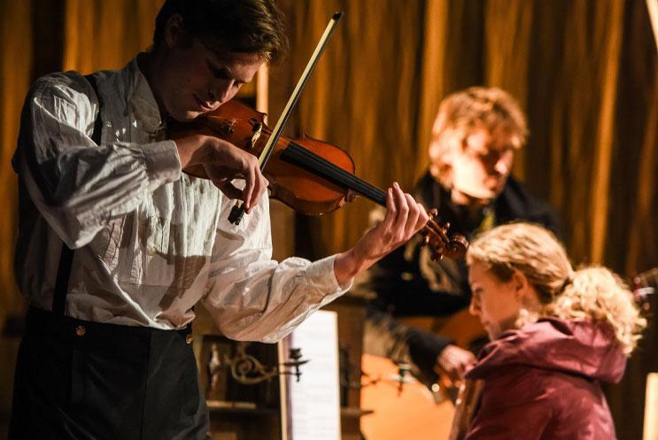 Characters in period drama costume. One is playing the violin.