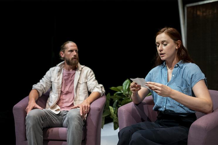 Two performers sat in chairs, one is reading a book.