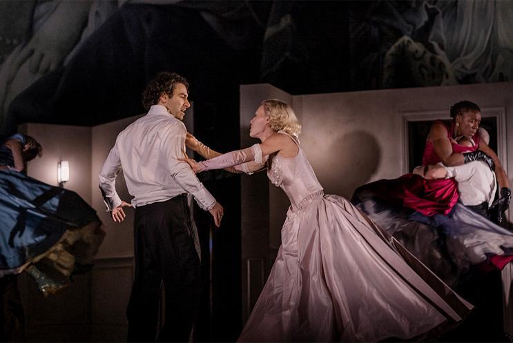 Two performers, both in old dinner party attire, clutching at each other in a heated exchange.