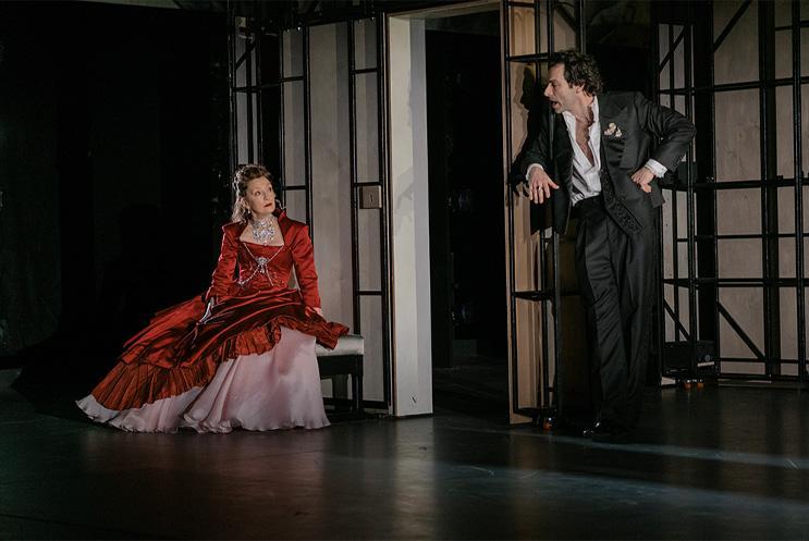 Two performers on stage talking too each other, one dressed in a suit, the other a dress.
