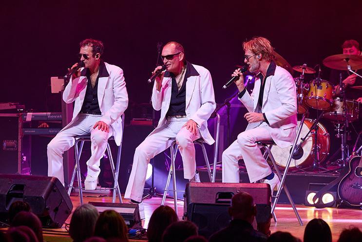 Three performers in white suits sat on stalls singing