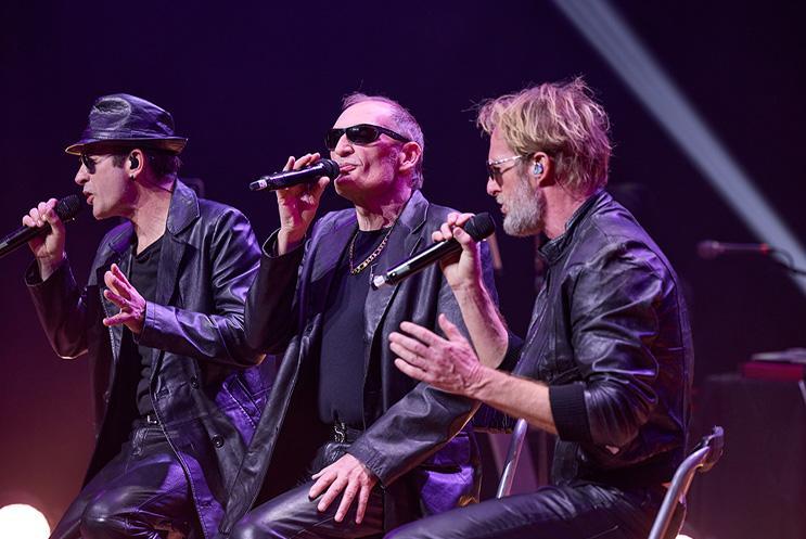 Three performers in black leather jackets singing into mics