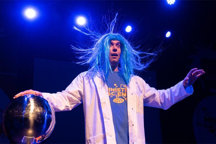 A person touching a magnetic ball, dressed in a white lab coat  and their long blue hair standing on end.