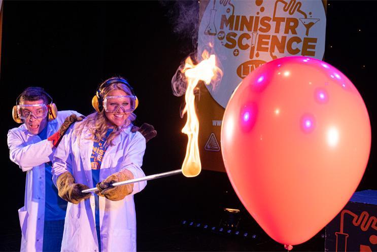 Two people in lab coats holding a long rod with flames at one end, pointing it at a big red balloon.