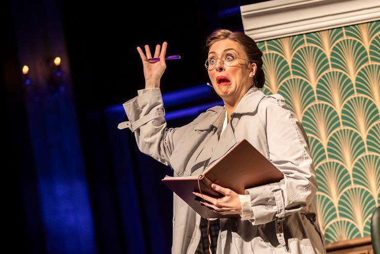 Woman wearing glasses and trench coat looking panicked while holding a book.