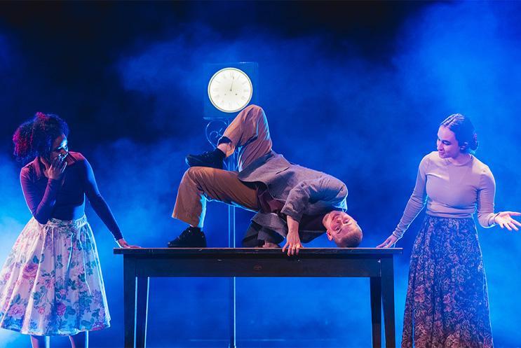 A performer bending into a distorted shape on top on a table with two other performers watching.