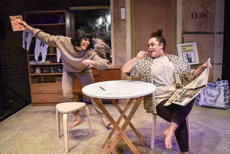 Two people around a kitchen table. One is sat laughing and the other is doing a dance