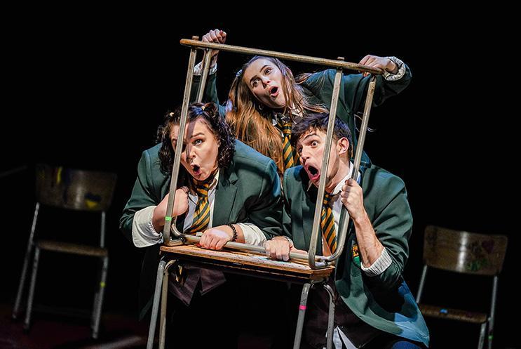 Three actors playing secondary school kids dressed in uniforms, peering through a box.