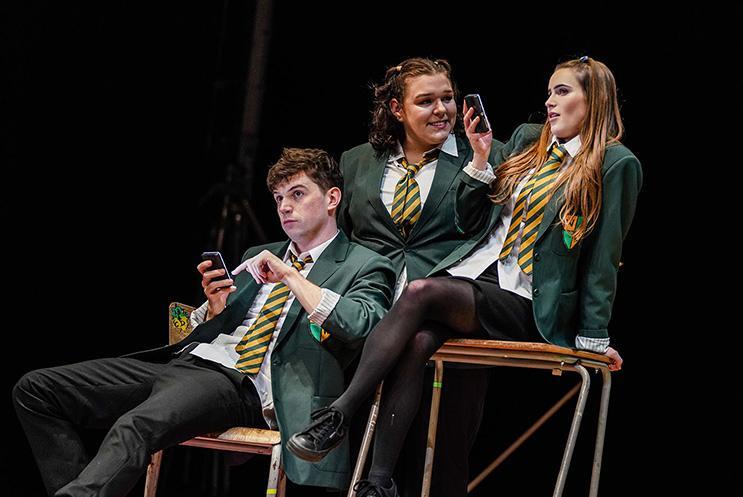 Three actors playing secondary school kids dressed in uniforms, on their phon