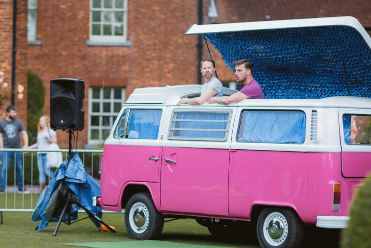 Two men poking their head out the top of a bright pink caravan