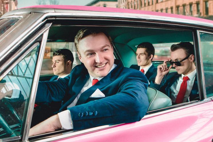 Four men in retro suits in a pink Cadillac